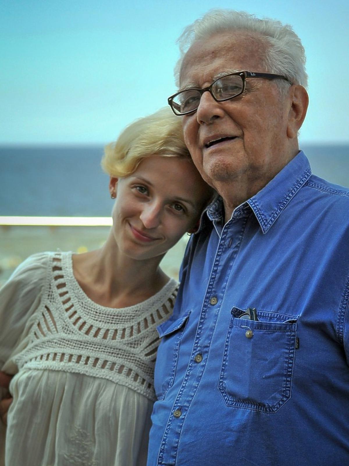 Kamila Łapicka w białej ażurowej bluzce stoi przytulona do męża Andrzeja Łapickiego w niebieskoej koszuli,w tle morze i plaża