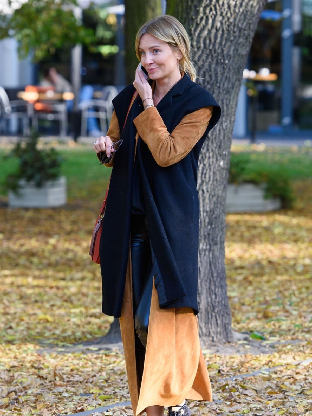 Agnieszka Szulim w zaszowym płaszczy, czarnej kamizelce, duzych okularach, czarne buty, czerwona torba