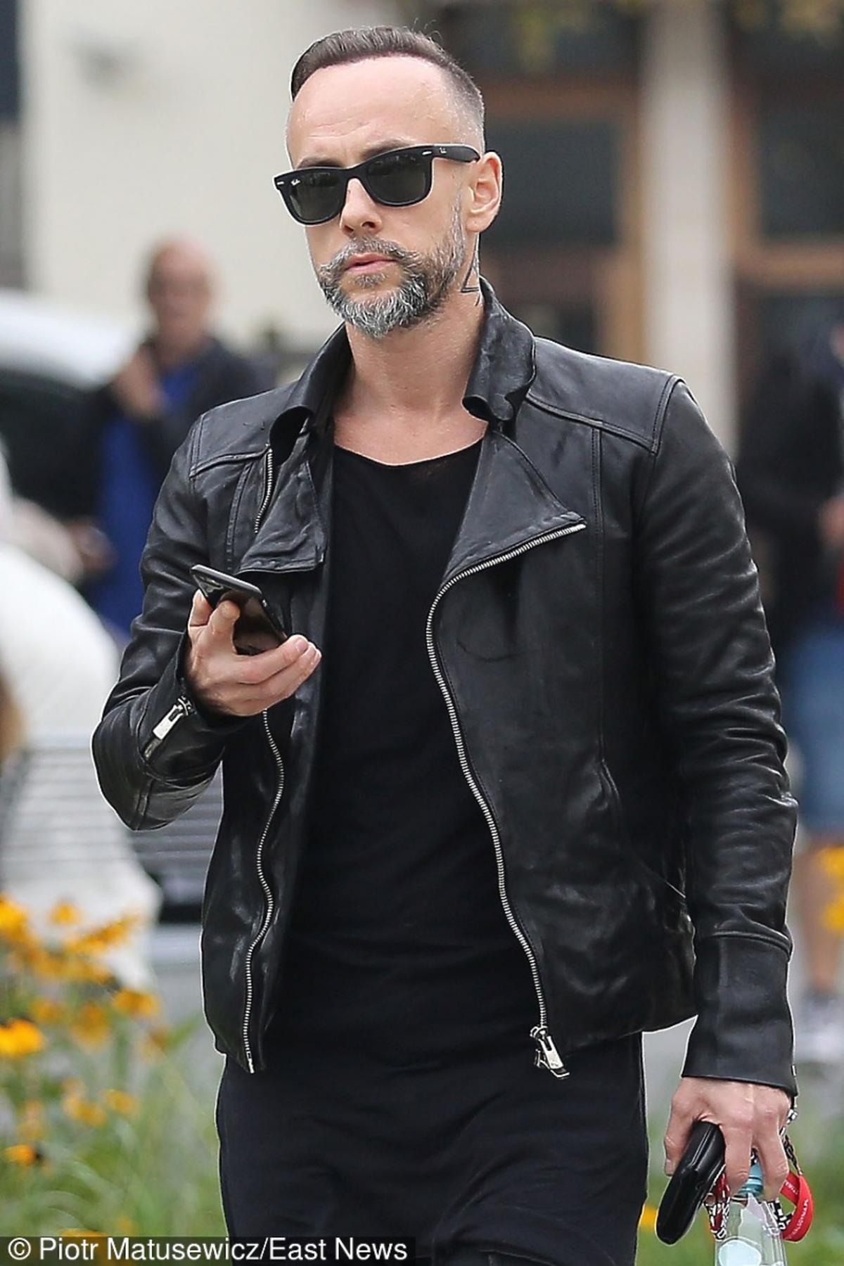 Nergal na ulicy w czarnej skórzanej kurtce, okularach przeciwsłonecznych trzyma w ręku telefon komórkowy