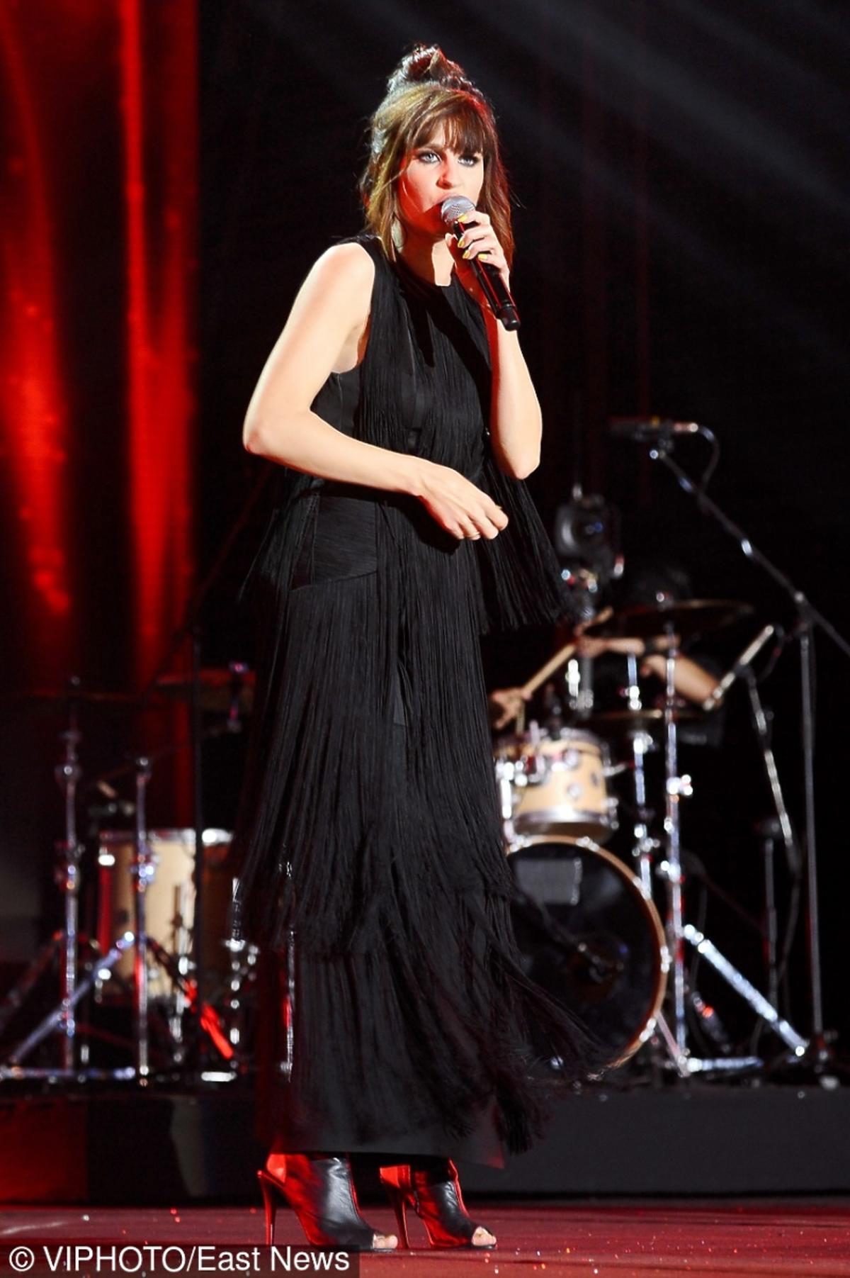 Sylwia Grzeszczak w czarnej długiej sukience, stoi na scenie z mikrofonem