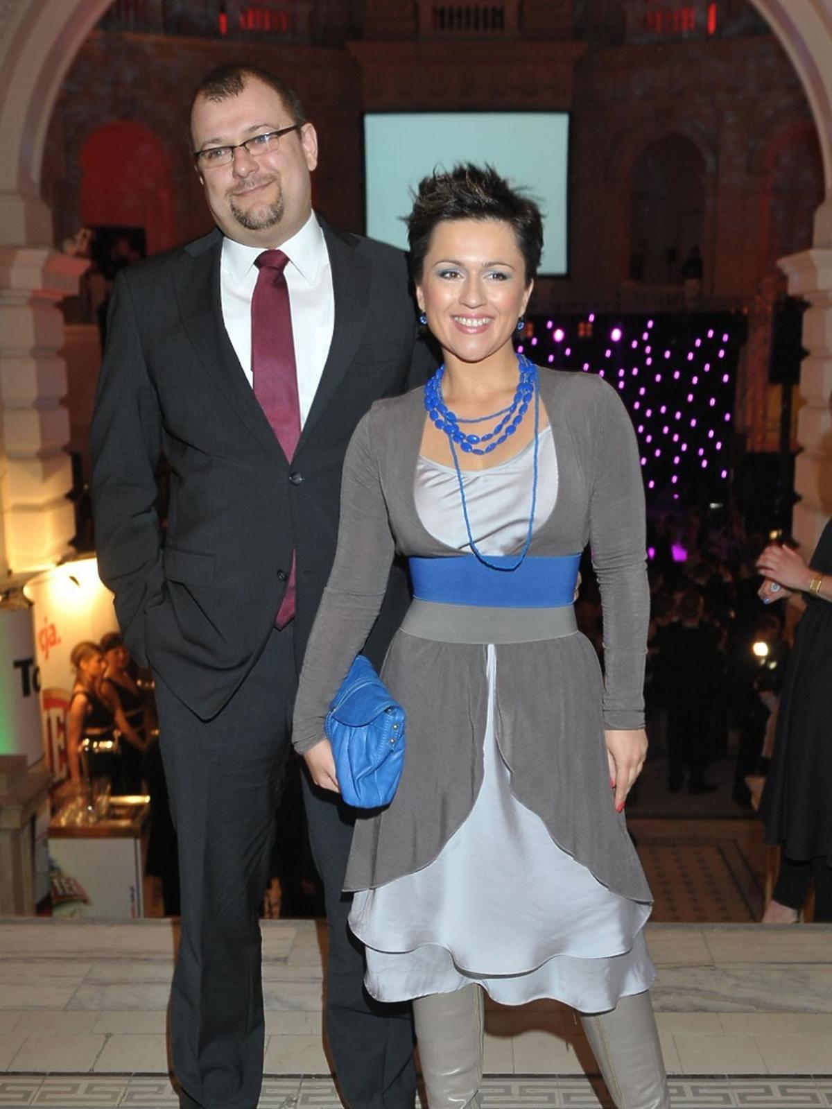 Beata Tadla w szarej sukience z niebieskimi dodatkami pozuje z ówczesnym mężem fotoreporterom podczas Charytatywnego Balu Dziennikarzy w auli Politechniki warszawskiej