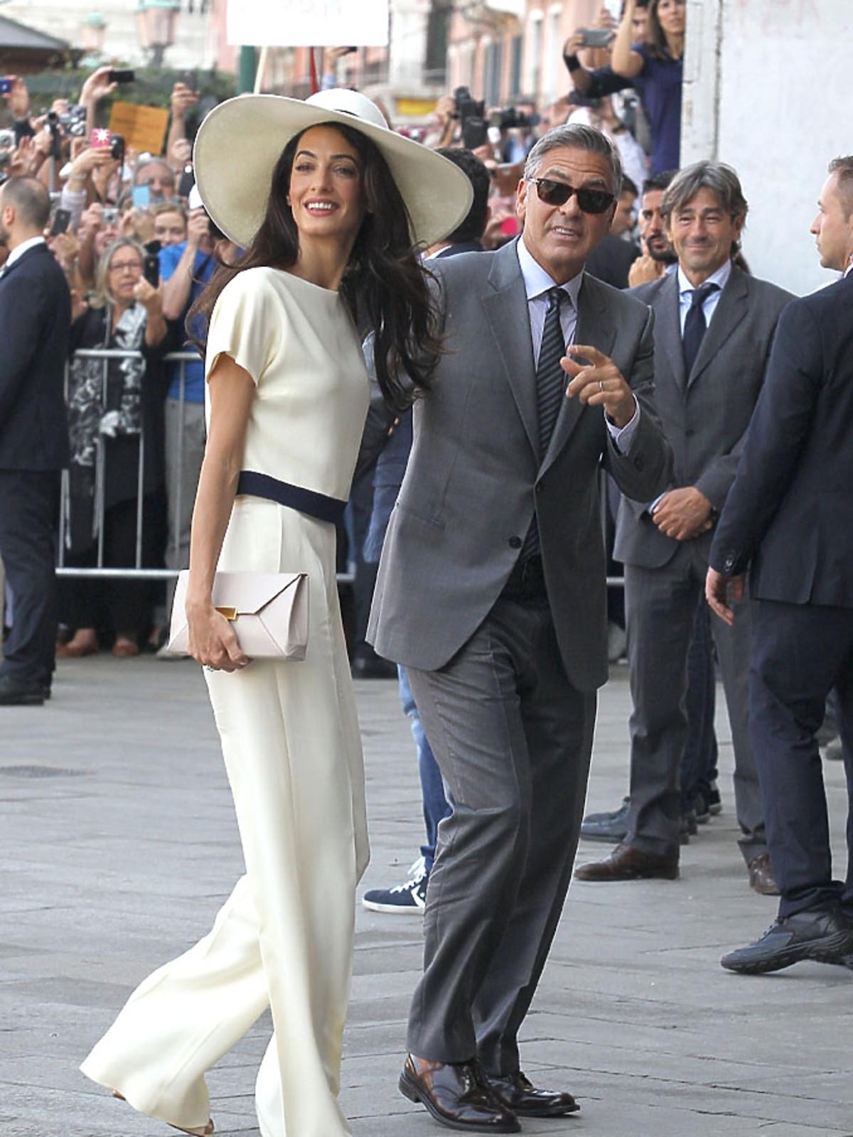 Amal Clooney ubrana na biało, w kapeluszu, z kopertówką, George Clooney w eleganckim grafitowym garniturze