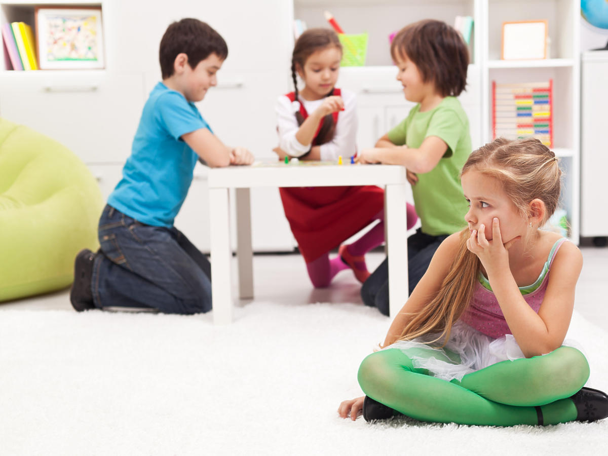 Dziewczynka siedząca na podłodze w przedszkolu, w tle inne dzieci.