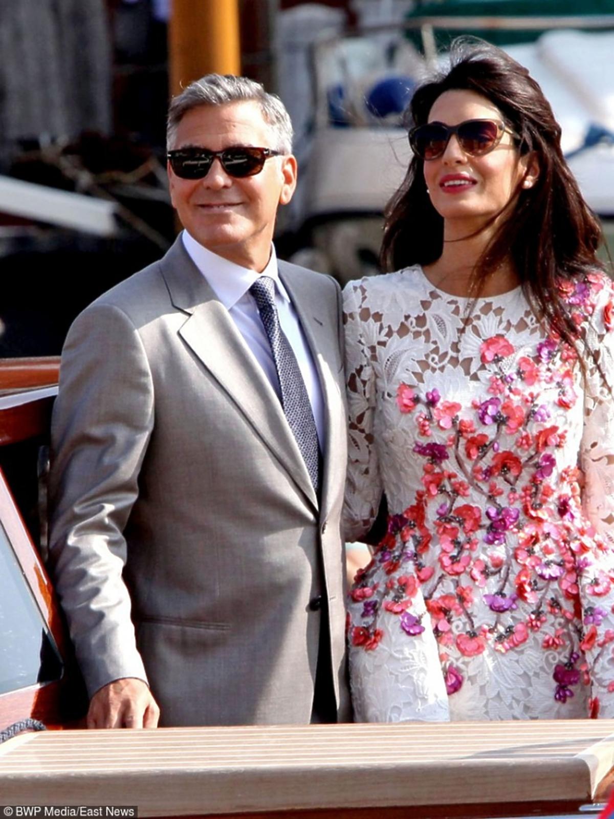 George Clooney w eleganckim grafitowym garniturze, Amal Clooney w białej koronkowej sukni z kolorowymi motywami kwiatowymi