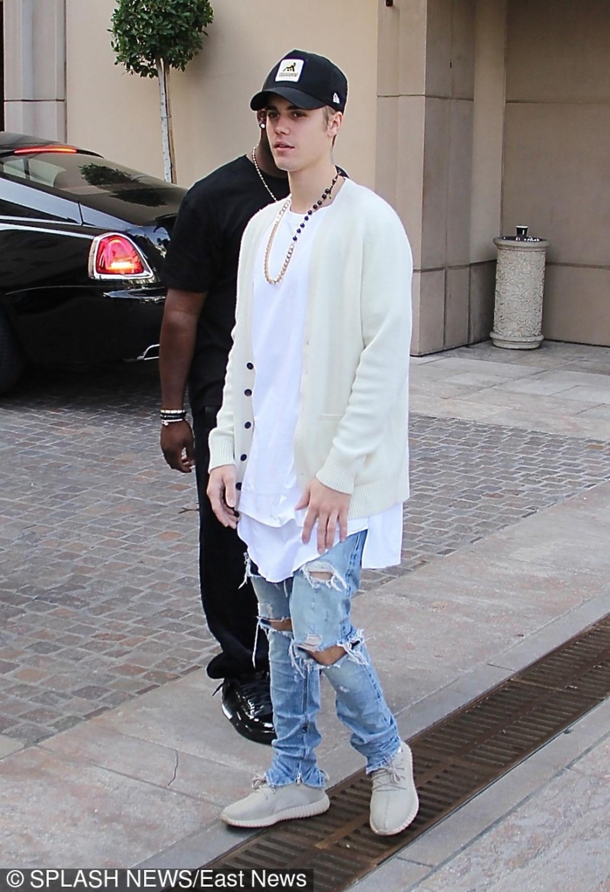 Justin Bieber w białej koszulce, czapce z daszkiem, podartych dzinsach, jasnych sportowych butach
