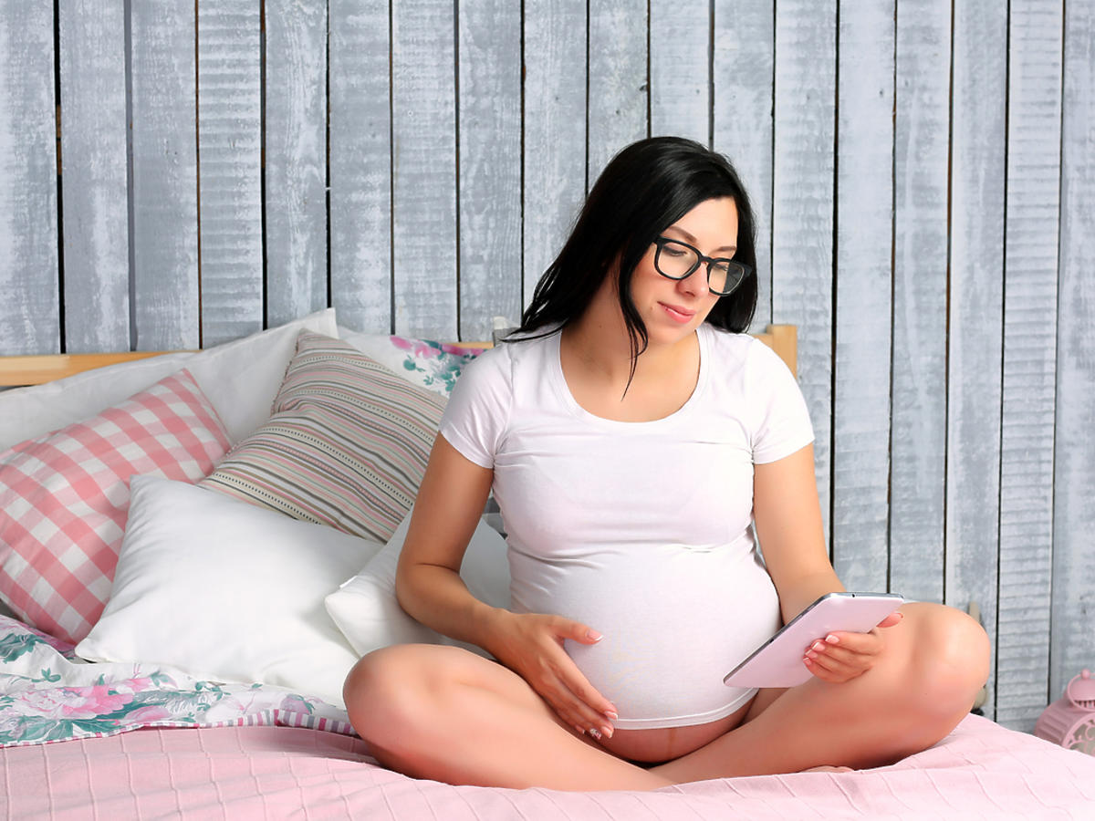 Kobieta w ciąży siedzi na łóżku i czyta coś na tablecie