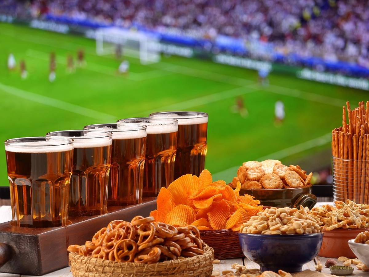 Krakersy, paluszki, piwo i orzeszki stoją na stole, w tle mecz w telewizji.