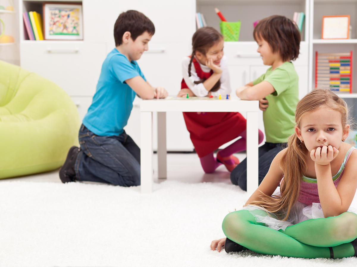 Mała dziewczyna siedzi na ziemi, w tle bawi się trójka innych przedszkolaków.