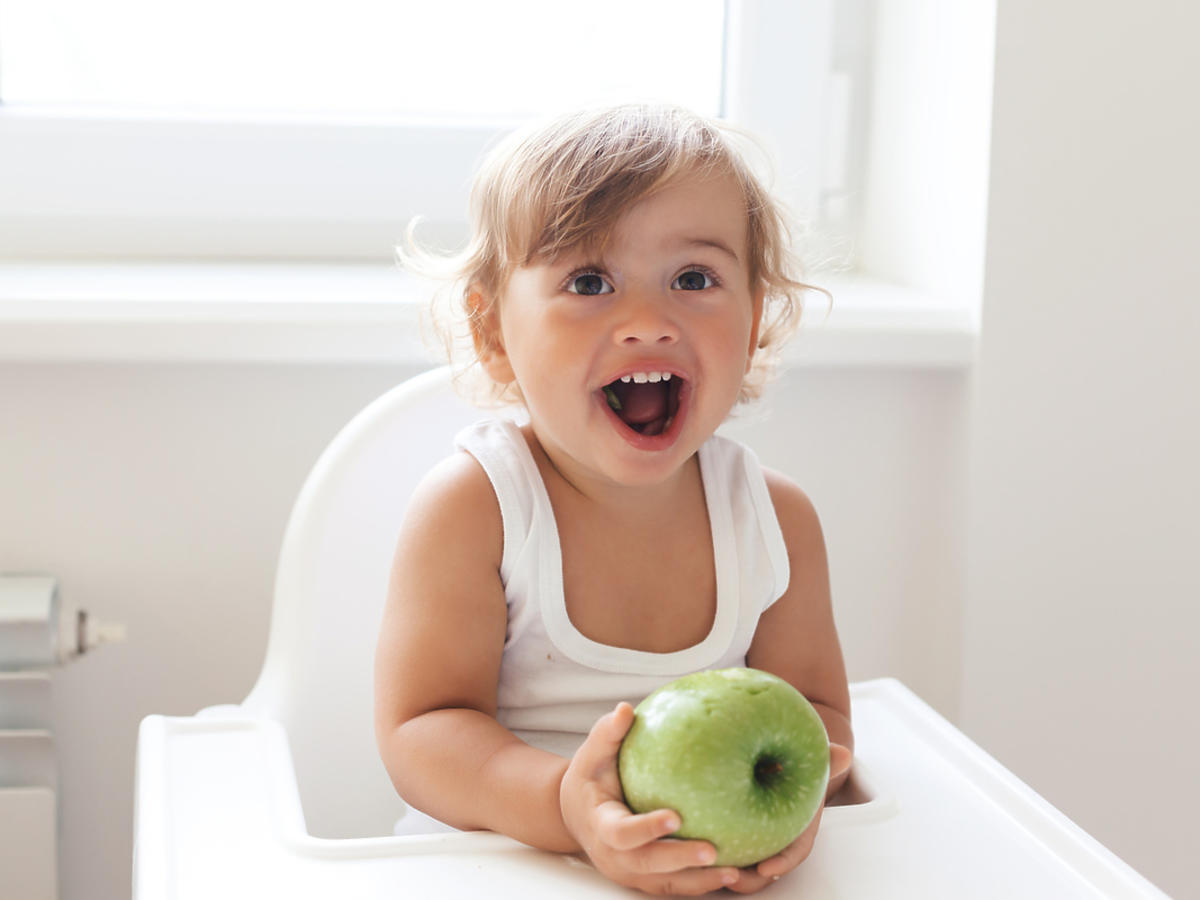 Małe dziecko siedzi w krzesełku do karmienia i trzyma w dłoniach jabłko.