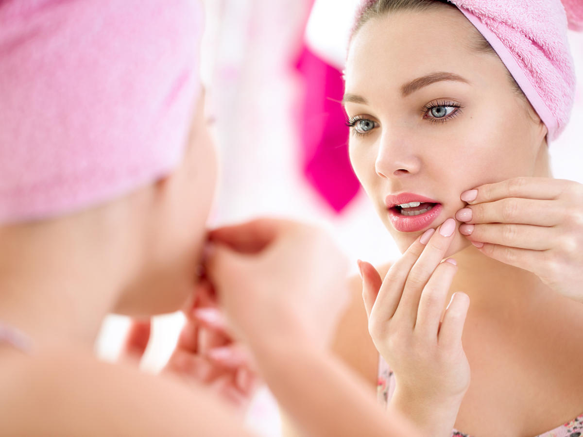Młoda kobieta przegląda się w lustrze i nakłada na twarz kosmetyk przeciw pryszczom.