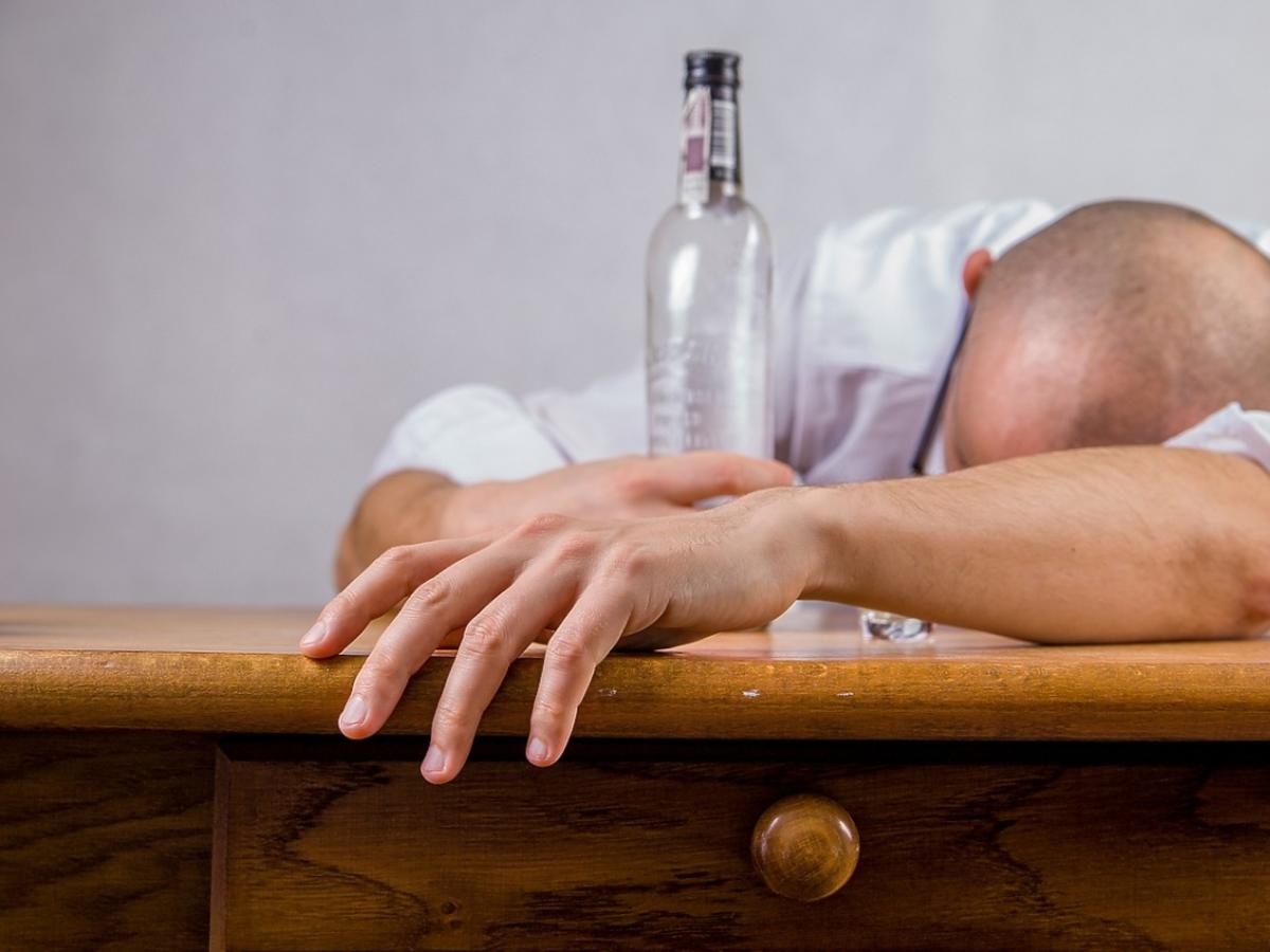 Pijany mężczyzna śpi na stole. W dłoni trzyma butelkę wódki.