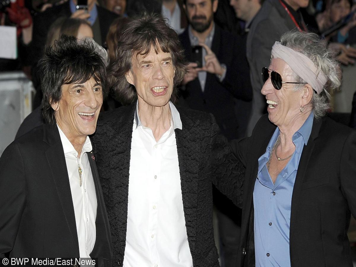 Ronnie Wood, Mick Jagger i Keith Richards w czarnych marynarkach obejmują się i śmieją